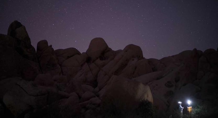 Two people wearing headlamps appear in the bottom right of the photo. Behind them, large boulders are illuminated in front of a purple starry sky.